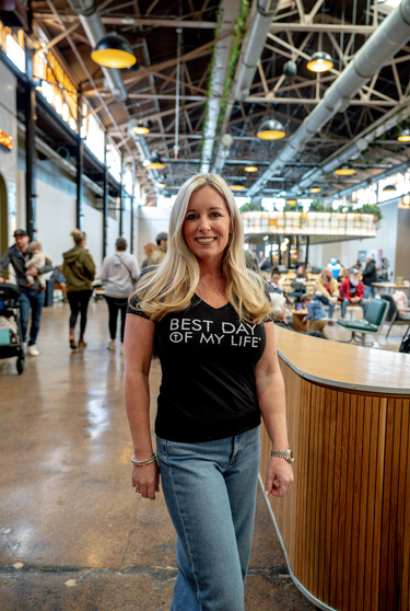 light-skinned person with long blonde hair wearing a black vneck t-shirt with best day of my life with a cross in an O written across in white block lettering.